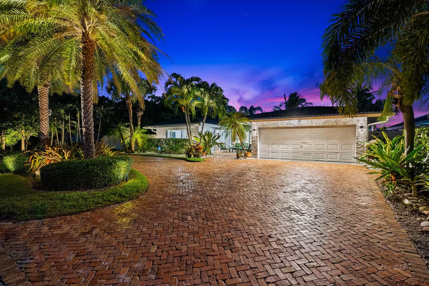 Exterior of luxury Pompano Beach vacation rental at night — brick paver driveway, tropical landscaping, double garage